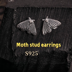 Op de afbeelding: Een paar zilverkleurige mot-oorstekers. De oorbellen zijn gedetailleerd met vleugel- en lichaamskenmerken. De tekst "Moth stud earrings" en "S925" is in gele letters.