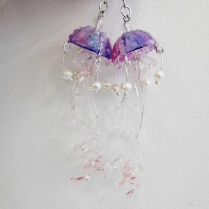 May include: A pair of jellyfish earrings. The earrings have pink and purple dome-shaped tops, with clear tendrils and faux pearl accents. The earrings are suspended from silver-coloured hooks. A unique piece of jewellery.