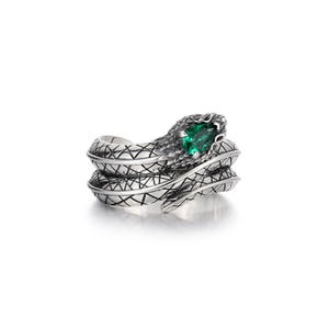Puede incluir: Un anillo de tono plateado con forma de serpiente enroscada que presenta un patrón de escamas texturizado y entrecruzado a lo largo del cuerpo. La cabeza de la serpiente está elevada y tiene una gema verde vibrante en forma de pera.