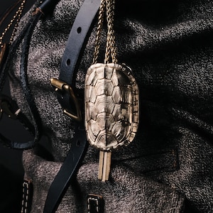 May include: A gold-colored turtle pendant with a detailed shell design hangs from a delicate chain. The pendant is suspended against a textured gray fabric and a black leather strap with a buckle. The pendant's intricate pattern and metallic sheen are highlighted.