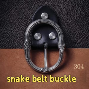 Op de afbeelding: Zilverkleurige slangenriemgesp met een zwarte leren achterkant. De gesp heeft een gedetailleerd slangenontwerp en een metalen pin. De tekst "snake belt buckle" staat in het geel onderaan en het nummer "304" rechtsboven.