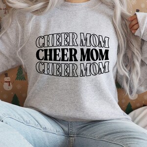 May include: A light gray sweatshirt with the words "CHEER MOM" stacked three times in black, outlined letters. The sweatshirt is made of a soft material and has long sleeves. The background is a festive pattern with snowmen and Christmas trees.