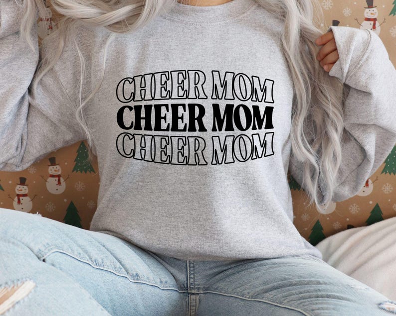 May include: A light gray sweatshirt with the words "CHEER MOM" stacked three times in black, outlined letters. The sweatshirt is made of a soft material and has long sleeves. The background is a festive pattern with snowmen and Christmas trees.
