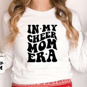 May include: White long-sleeve shirt with the text "IN MY CHEER MOM ERA" in a black, retro font. A white mug with "Cheer Mom" printed on it is also visible. The shirt is a casual style.