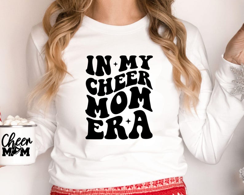 May include: White long-sleeve shirt with the text "IN MY CHEER MOM ERA" in a black, retro font. A white mug with "Cheer Mom" printed on it is also visible. The shirt is a casual style.