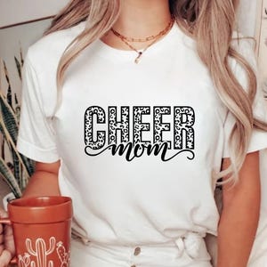 May include: White t-shirt with the words "CHEER MOM" in a black, leopard print design. The shirt is short-sleeved and has a crew neck. A person is holding a brown mug with a cactus design.