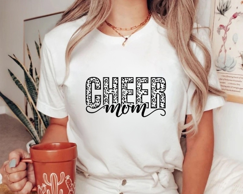May include: White t-shirt with the words "CHEER MOM" in a black, leopard print design. The shirt is short-sleeved and has a crew neck. A person is holding a brown mug with a cactus design.