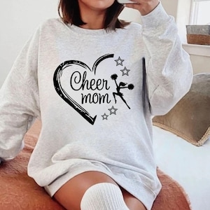 May include: A light gray sweatshirt with a black heart design that says "Cheer mom." The heart has a cheerleader silhouette and stars. The sweatshirt is long-sleeved and has a relaxed fit.