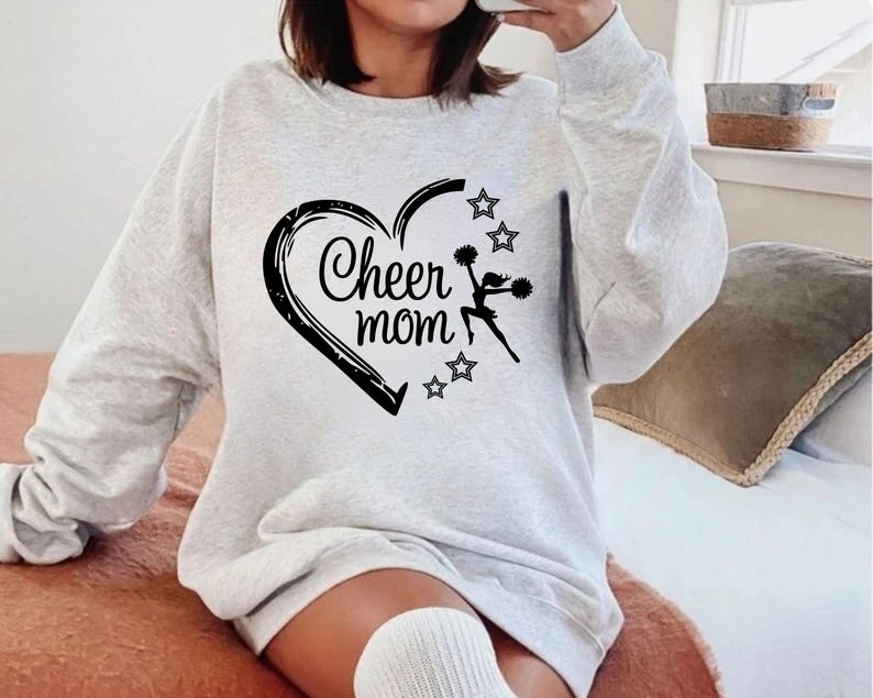 May include: A light gray sweatshirt with a black heart design that says "Cheer mom." The heart has a cheerleader silhouette and stars. The sweatshirt is long-sleeved and has a relaxed fit.
