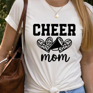 May include: Cream-colored t-shirt with the words "CHEER mom" in black block letters and script. The design includes a megaphone and two leopard print hearts. The shirt is a casual, everyday wear item.
