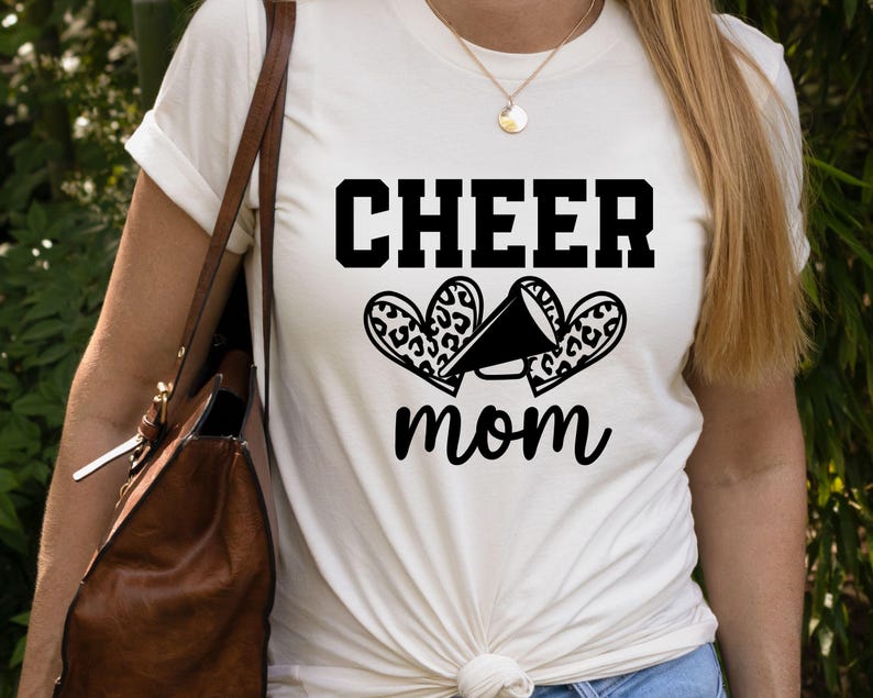 May include: Cream-colored t-shirt with the words "CHEER mom" in black block letters and script. The design includes a megaphone and two leopard print hearts. The shirt is a casual, everyday wear item.