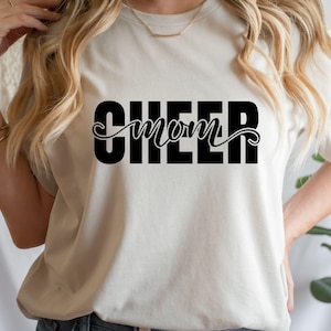 May include: Cream-colored t-shirt with the words "CHEER mom" printed in black. The word "CHEER" is in a bold, block font, while "mom" is in a cursive script. The shirt is a casual, everyday wear item.