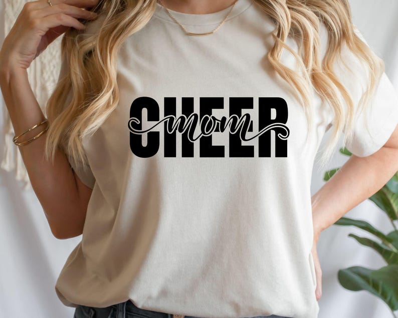 May include: Cream-colored t-shirt with the words "CHEER mom" printed in black. The word "CHEER" is in a bold, block font, while "mom" is in a cursive script. The shirt is a casual, everyday wear item.