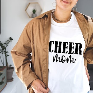 May include: White t-shirt with the text "CHEER mom" in black, worn under a brown button-down shirt. The person is wearing a gold chain necklace and hoop earrings. The background includes plants and a television.