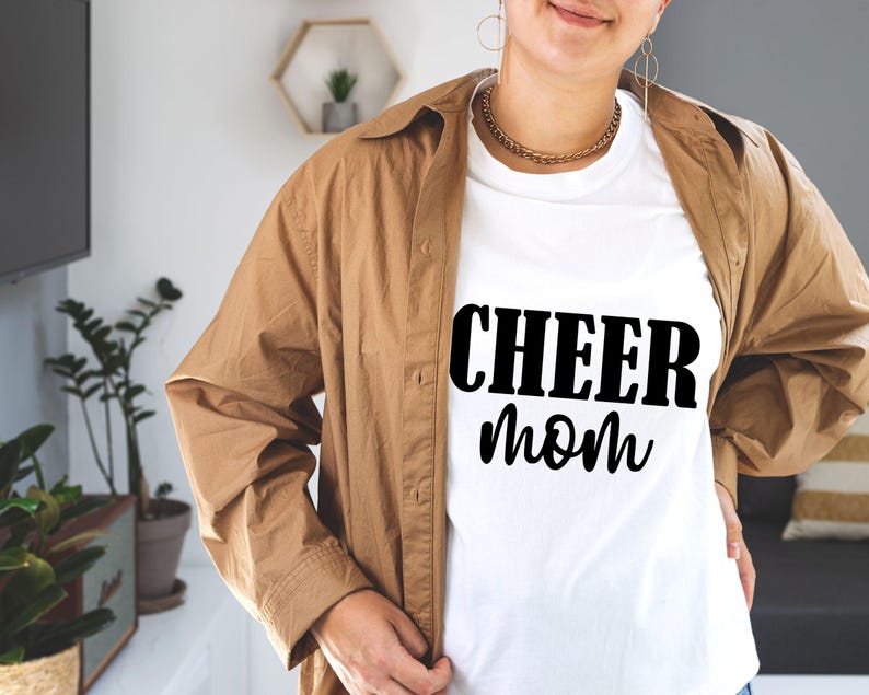 May include: White t-shirt with the text "CHEER mom" in black, worn under a brown button-down shirt. The person is wearing a gold chain necklace and hoop earrings. The background includes plants and a television.