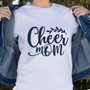 May include: White crew neck sweatshirt with the navy blue text "Cheer Mom" in a cursive font, with a decorative leaf design above the word "mom". The sweatshirt is being worn with a blue denim jacket.