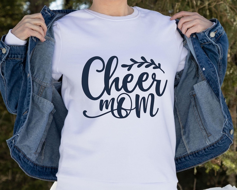 May include: White crew neck sweatshirt with the navy blue text "Cheer Mom" in a cursive font, with a decorative leaf design above the word "mom". The sweatshirt is being worn with a blue denim jacket.
