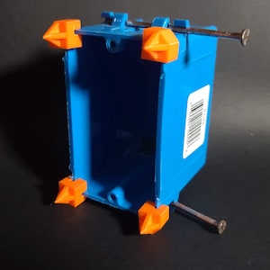 Puede incluir: Una caja eléctrica azul con soportes de montaje naranjas y clavos. La caja tiene forma rectangular con un interior abierto. Una etiqueta de código de barras es visible. Diseñada para cableado eléctrico.