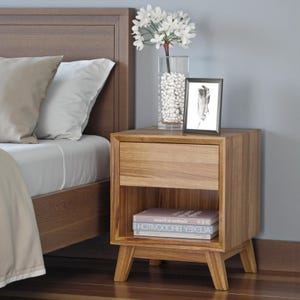 May include: A wooden bedside table with a drawer and an open shelf. The table has a natural wood finish and sits on angled legs. A vase of white flowers, a framed picture, and books are on top. The bed frame is visible.