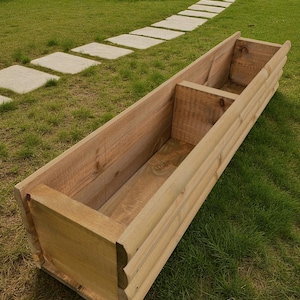May include: A rectangular wooden planter box with a natural finish. The box is divided into two sections and sits on a grassy lawn. The planter has a decorative edge and is ready for planting flowers or herbs.