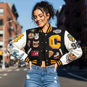 May include: Black varsity jacket with white sleeves, adorned with patches like a peace symbol, a cat wearing sunglasses, and a pizza slice. The jacket has yellow and black striped trim and a large letter "C" on the chest.