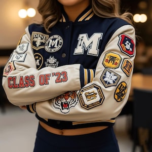 Może przedstawiać: Granatowa i kremowa kurtka typu letterman z wieloma naszywkami. Kurtka ma literę "M" na piersi i napis "CLASS OF '23" na rękawie. Naszywki obejmują tygrysa, różne emblematy i tekst.