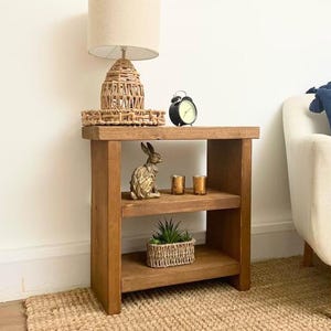 May include: A two-tiered wooden side table with a beige lamp, black alarm clock, gold rabbit ornament, two gold candle holders, and a small plant in a woven basket. The table is on a woven rug. The table is made of wood.