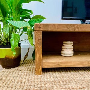 May include: A rustic, wooden TV stand with a lower shelf. A stack of three decorative, white and gold-banded objects sits on the shelf. A potted plant with large green leaves is to the left, and a TV is in the background.