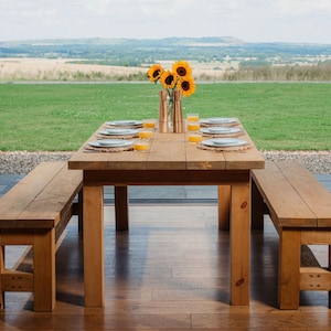 May include: A wooden outdoor dining set with a rectangular table and two benches. The table is set with plates, placemats, and glasses of juice. A vase of sunflowers sits in the center. The table is approximately 183 cm long.