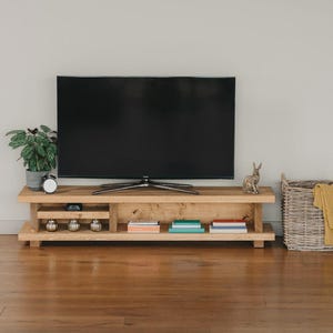 May include: A natural wood TV unit with a large black screen. The unit features a lower shelf with books and decorative items. A plant, clock, and rabbit ornament are also on display.