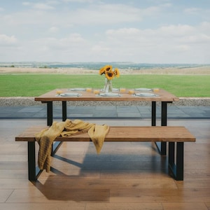 May include: A rectangular wooden dining table and a matching wooden bench, both featuring black metal U-shaped legs. A clear glass vase with yellow sunflowers sits in the centre of the table, which is set with white plates and orange glasses.