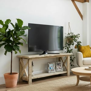 May include: A light-coloured wooden TV stand with an X-shaped design on the sides. A large black television sits on top. Two framed photos are on the lower shelf. A potted plant and a hanging plant are also visible.