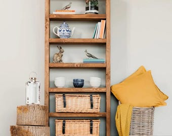 May include: A wooden bookshelf filled with decorative items. The shelves hold a teapot, books, bowls, and figurines. Wicker baskets with black straps are on the lower shelves. A lantern and tree stumps are on the left, and a basket with pillows is on the right.