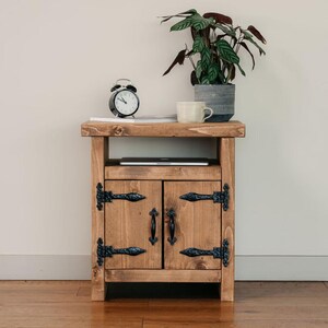 May include: A wooden bedside cabinet with two doors, black hinges, and handles. The cabinet has a shelf and is topped with a black alarm clock, a mug, a potted plant, and a laptop. The wood has a warm, natural finish. The cabinet is approximately 60cm tall.