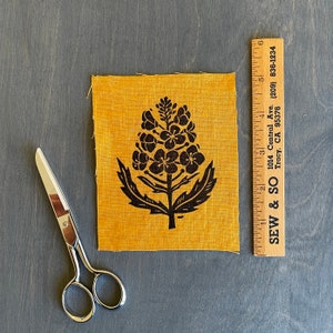 Könnte beinhalten: Ein gelber Stoff-Patch mit einem schwarzen Blumenmuster. Das Design zeigt eine stilisierte Blume mit mehreren Blütenblättern und Blättern.