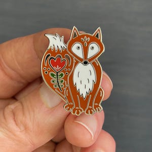 May include: A decorative fox pin with a folk art design. The pin features a brown fox with white fur on its chest and a stylized tail with a red flower and green leaves. The pin is outlined in gold.