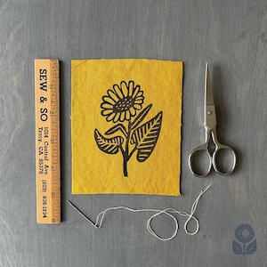 May include: A yellow fabric square with a black sunflower design, a wooden ruler with "SEW & SO" text, scissors, a needle, and white thread. The ruler measures up to 6 inches. The image is on a gray surface.