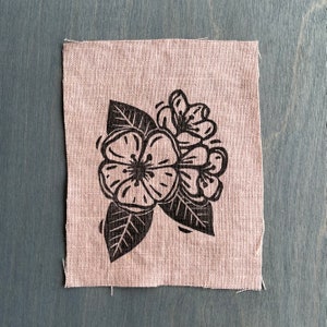 May include: A black and white floral design printed on a beige fabric patch. The design features a large flower with five petals and a smaller flower with four petals.
