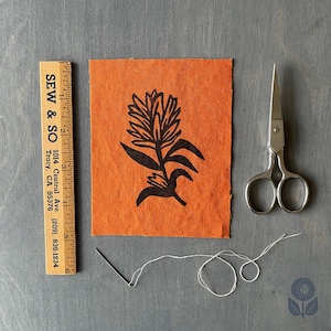 May include: An orange fabric square with a black floral design, a wooden ruler, scissors, a needle, and white thread. The ruler reads "SEW & SO" and includes a phone number and address. The scissors are silver and the needle is thin.