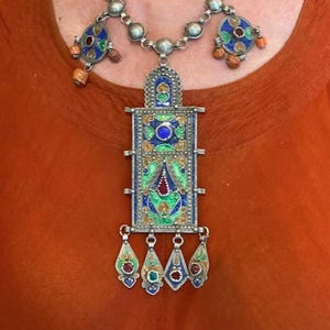 May include: A silver necklace with a large, rectangular pendant featuring intricate geometric designs in blue, green, red, and gold enamel. The pendant has four smaller, teardrop-shaped pendants hanging from it.