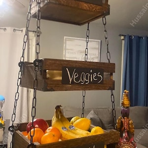 May include: A three-tiered hanging wooden fruit and vegetable basket. The top tier is empty. The middle tier has the word "Veggies" written on it. The bottom tier is filled with fruit and has the word "Fruit" written on it. The basket is suspended by chains.