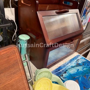May include: A wooden bread box with a clear glass lid. The bread box has a handle on the front and is sitting on a table with a blue and white patterned surface.