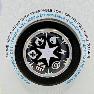 May include: A black and white phone grip with a star and various symbols. The grip is on a white card with the text "POPSOCKETS" and "POPGRIP" in colorful letters. The card also has text in French and English about the phone grip's features.