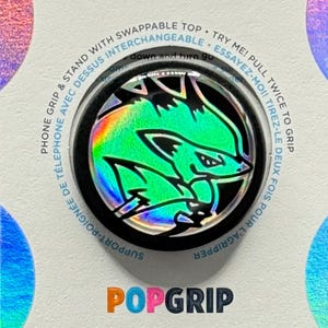 May include: A PopSockets PopGrip phone grip and stand with a swappable top. The grip features a black base with a holographic design of a green and blue creature. The packaging is white with the PopSockets logo.