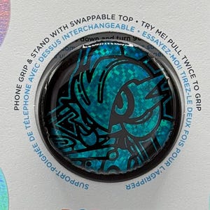 May include: A PopSockets PopGrip with a teal and black design. The packaging includes the PopSockets logo and text that reads "Phone grip & stand with swappable top". The PopGrip is designed to attach to the back of a phone.