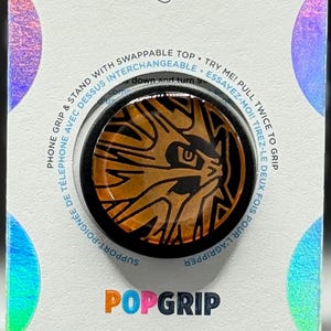 Op de afbeelding: Een PopSockets PopGrip met een zwart en oranje ontwerp. De verpakking bevat de logo's PopSockets en PopGrip, samen met tekst in het Engels en Frans. De PopGrip is ontworpen om aan een telefoon te bevestigen.
