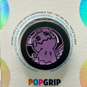 Könnte beinhalten: Ein PopSockets PopGrip Handyhalter und Griff in Einzelhandelsverpackung. Der runde Griff zeigt eine lila Mimikyu-Charaktergrafik. Die weiße Trägerkarte enthält den Text PopSockets, PopGrip sowie Gebrauchsanweisungen in Englisch und Französisch.
