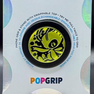 May include: A PopSockets PopGrip phone stand and grip in original packaging. The circular grip features a bright yellow and black graphic design. The white backing card displays the text POPSOCKETS and POPGRIP with holographic accents.