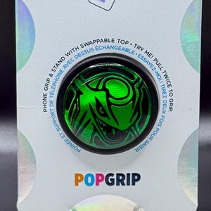 May include: A PopSockets PopGrip phone stand and grip in retail packaging. The circular grip features a green graphic of a creature. The white backing card displays the text PopSockets and PopGrip, with instructions to pull twice to grip.