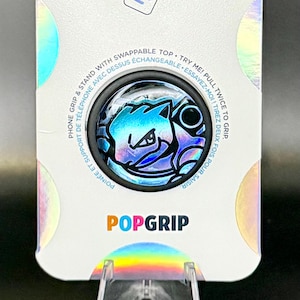 Puede incluir: Un PopGrip para teléfono azul y negro con acabado holográfico. El agarre presenta una imagen de dibujos animados de una criatura azul y negra con un ojo negro. El texto "PopGrip" está impreso en letras rosa, azul y verde en el embalaje.